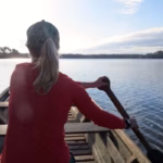 a person rowing a wooden canoe on a body of water, likely an oxbow lake in the Peruvian Amazon rainforest, such as Sandoval Lake puerto maldonado day tours